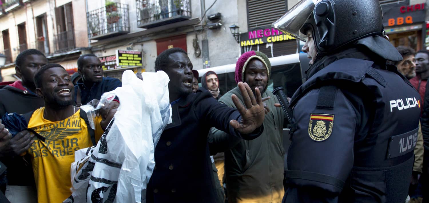 Grupo de personas negras en una calle de Madrid levantando las manos y bolsas frente a un agente de policía, durante una identificación que ilustra...