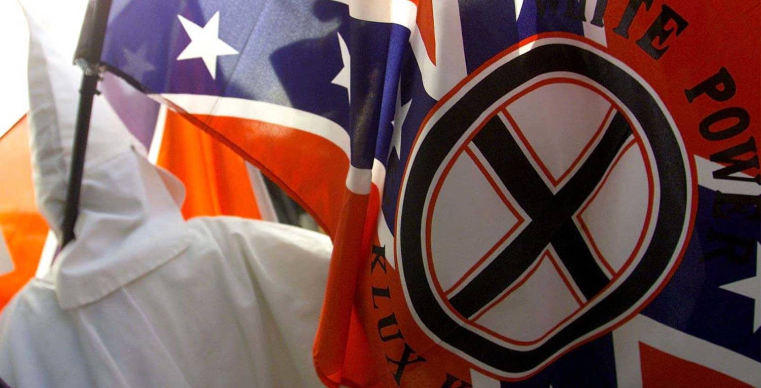 Miembro del Ku Klux Klan cubierto con túnica blanca ocultándose tras banderas en una protesta