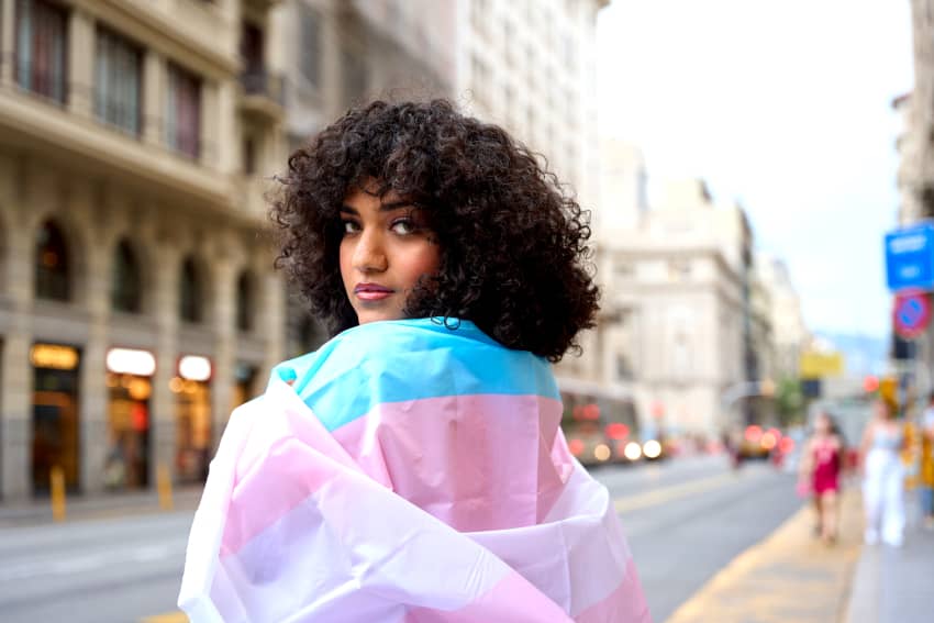 Una mujer con el pelo rizado y envuelta con una bandera trans está de pie en una calle mientras mira a cámara.