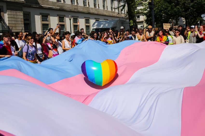 Manifestación del Orgullo con múltiples manifestantes sosteniendo una bandera trans de grandes dimensiones