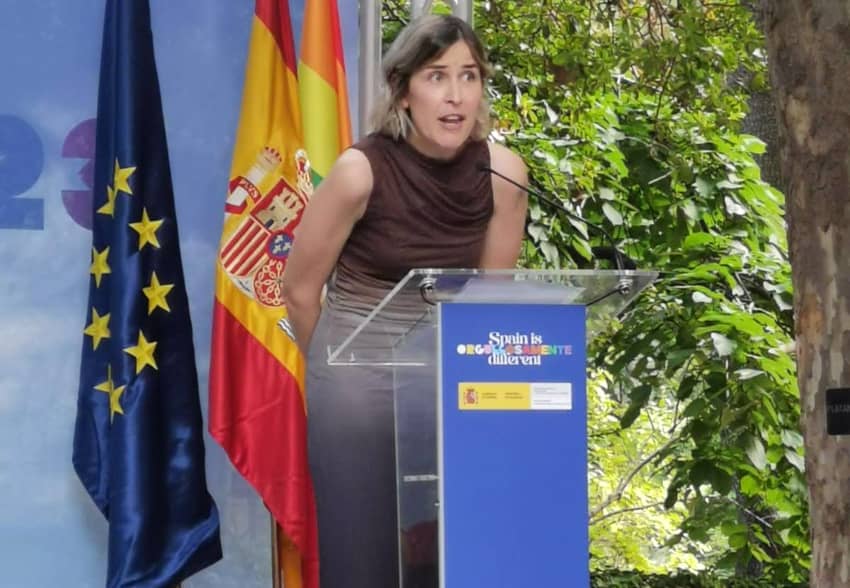 Camino Baró pronuncia un discurso en un acto público, con las banderas de España y la Unión Europea al fondo.